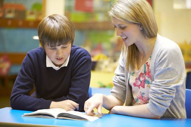 Teacher With Male Pupil Reading At Desk In Classroom